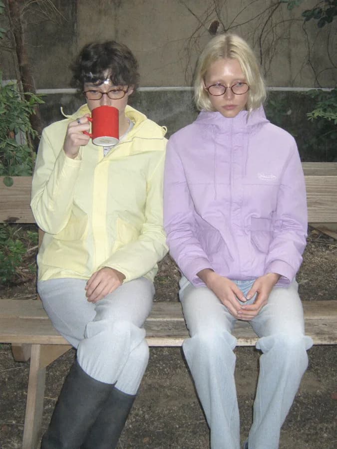Two young adults sitting side by side on a wooden bench at a worn picnic table in a small, plant-filled backyard, both w